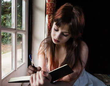 A young woman reflecting and writing in a notebook by the window, exploring Reflection Questions for Self-awareness.